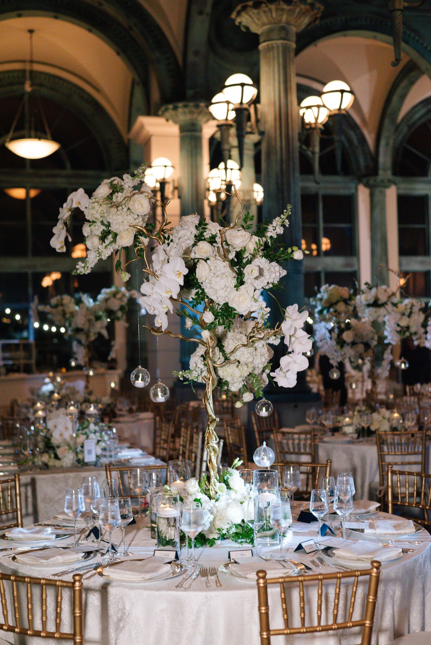 Tall white floral centerpiece with orchids and roses on gold chiavari chair reception table in grand ballroom.