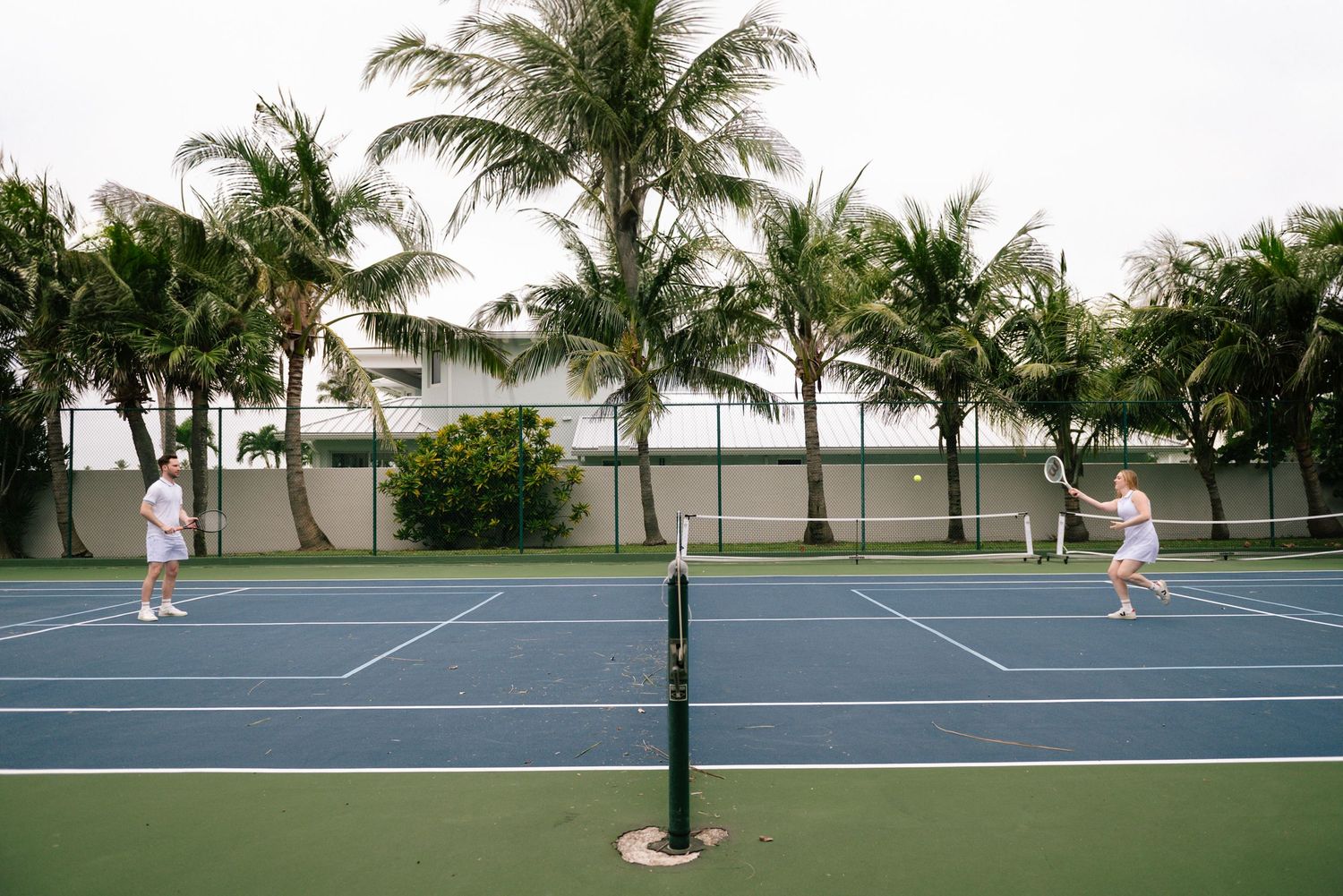 Players enjoy a tennis match on a blue court surrounded by tropical palm trees at a beachside resort.