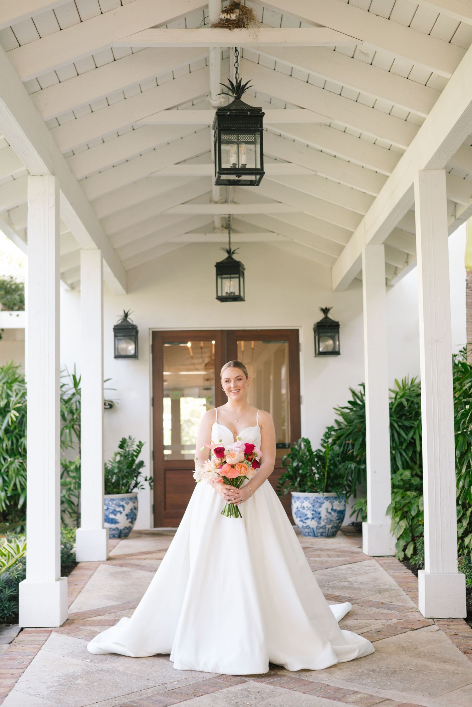 Bride in elegant white ball gown poses in covered walkway with tropical landscaping.