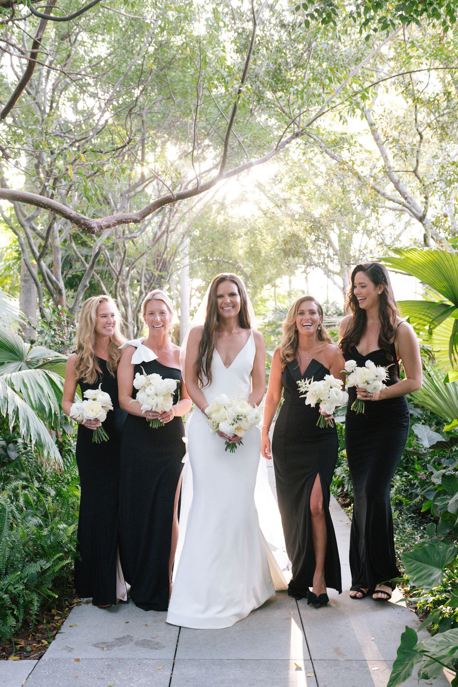Wedding party in black bridesmaid dresses and white bridal gown pose together in a lush garden setting.