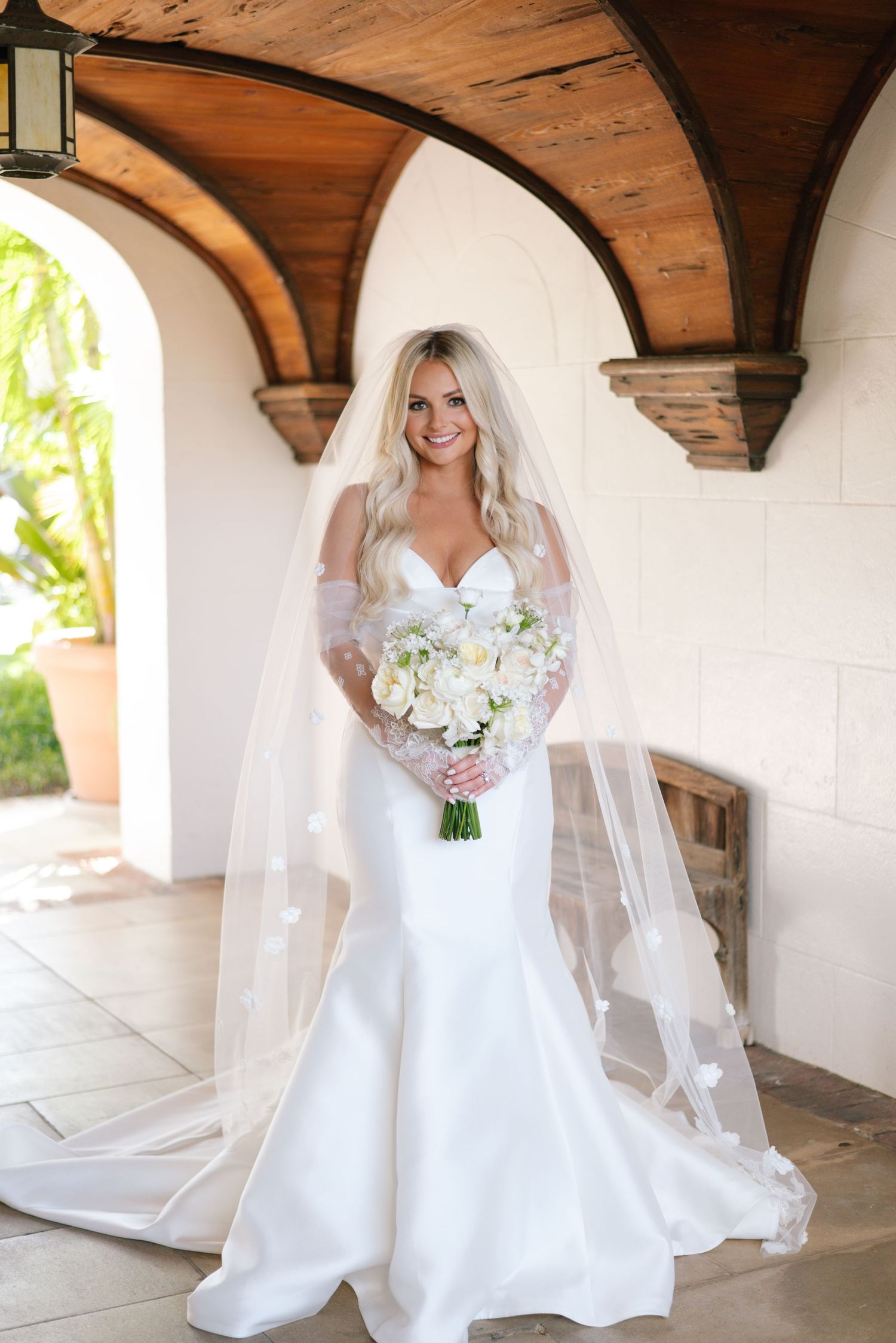 A bride in an elegant white wedding gown holds a bouquet of white flowers while standing in an arched corridor.