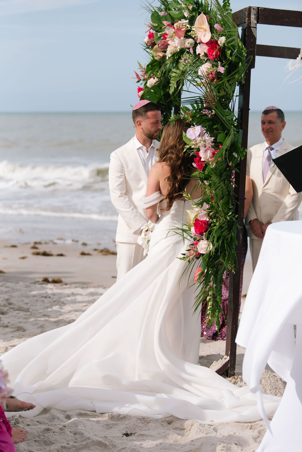 Beach wedding ceremony setup with floral arch and flowing white dress against ocean waves.