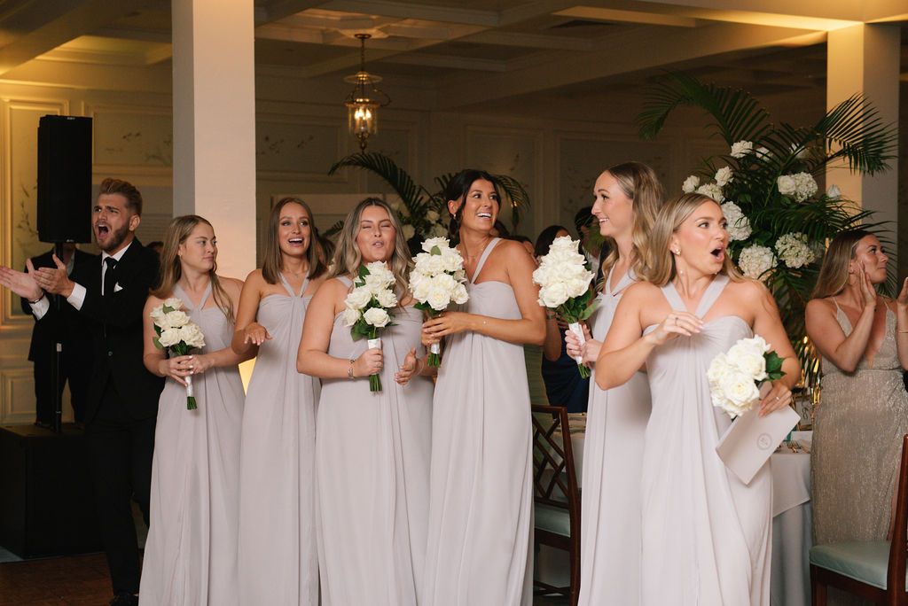 Wedding party in light colored dresses holding bouquets in an elegant reception hall.