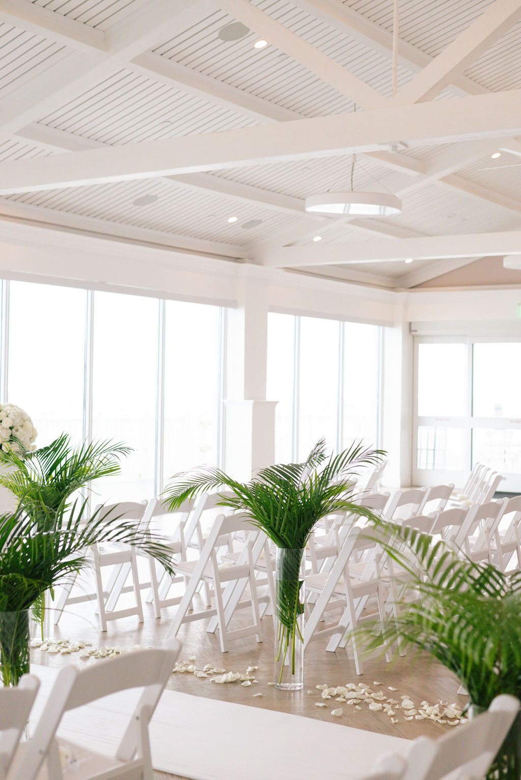 Modern white wedding ceremony space with rows of chairs and palm plants lining the aisle.