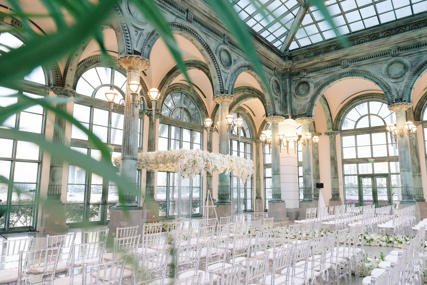 Wedding ceremony space with glass ceiling and rows of clear chairs in elegant ballroom.