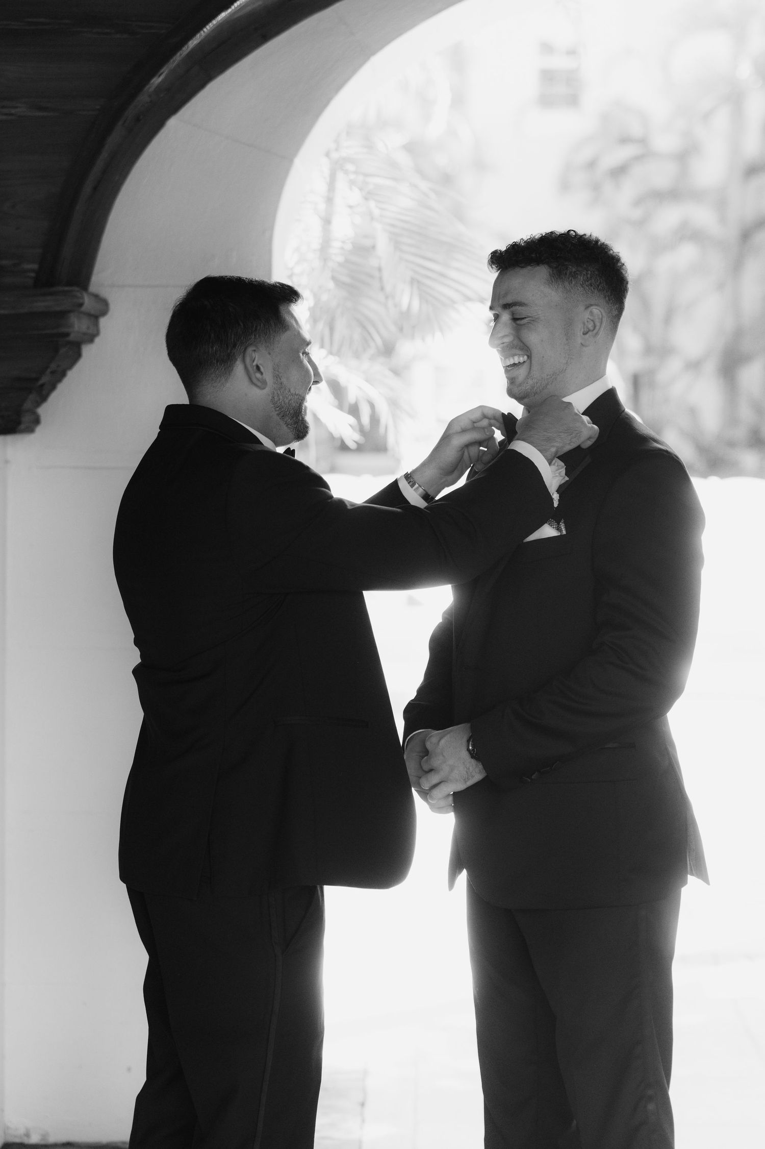 Two groomsmen in black tuxedos share a candid moment under an archway.