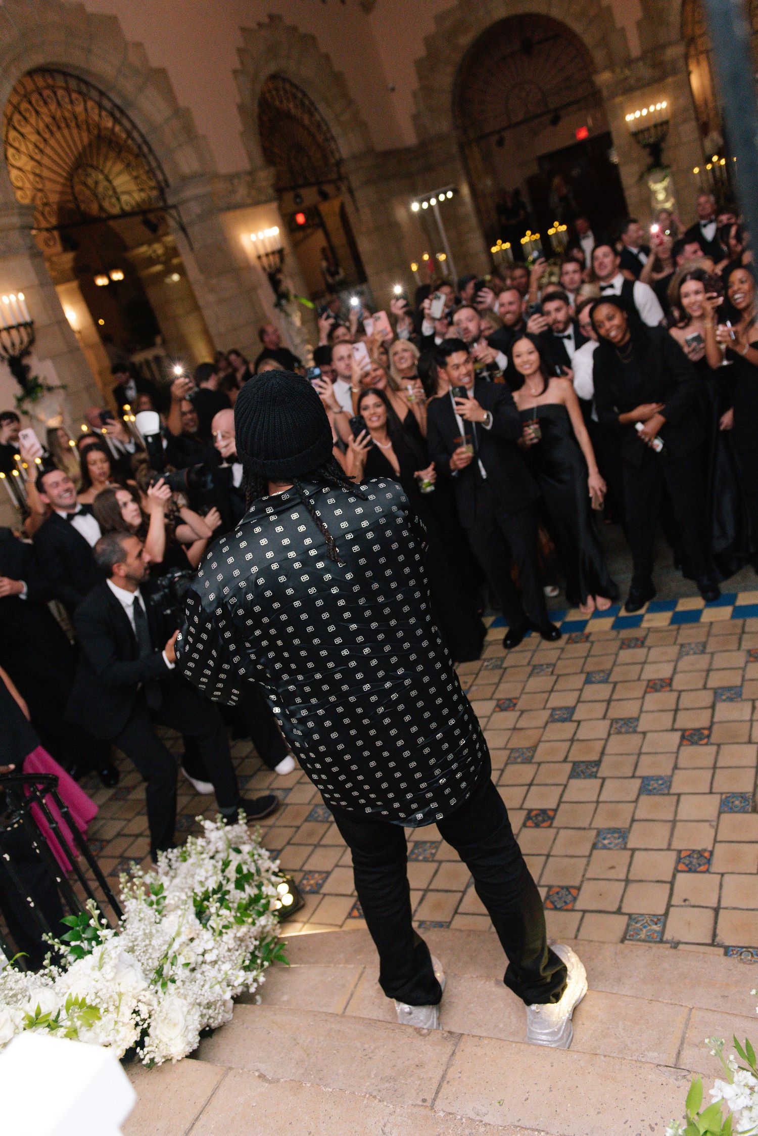 Wedding guests in formal attire watching performers dance on a tiled ballroom floor.