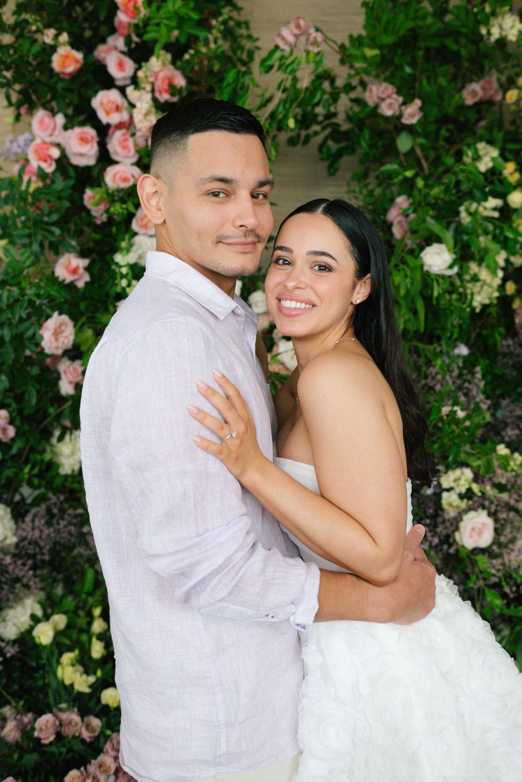 A romantic wedding photoshoot against a floral wall backdrop with pink roses and greenery.