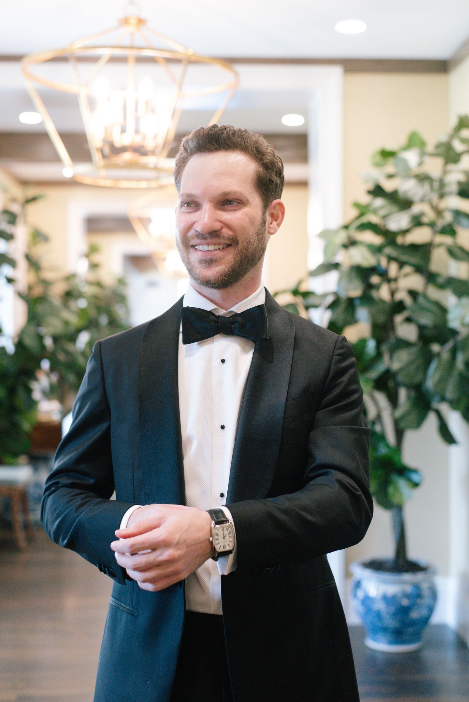 A formal event attendee wearing a classic black tuxedo and bow tie stands in an elegant venue with decorative lighting.