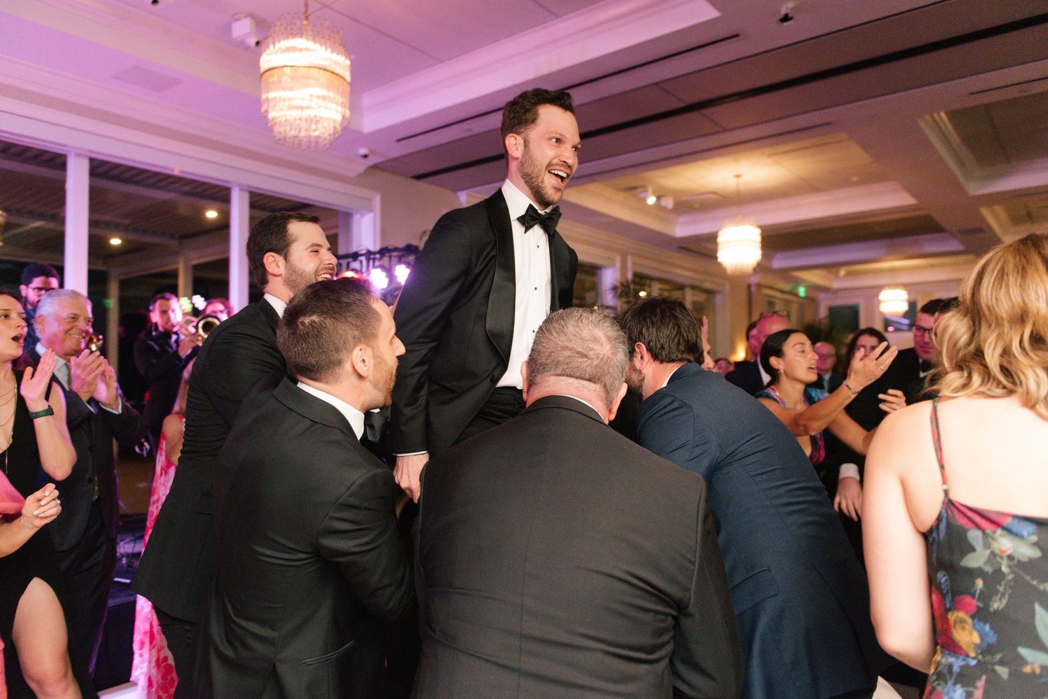 Wedding guests dancing and celebrating on a crowded dance floor under purple mood lighting.