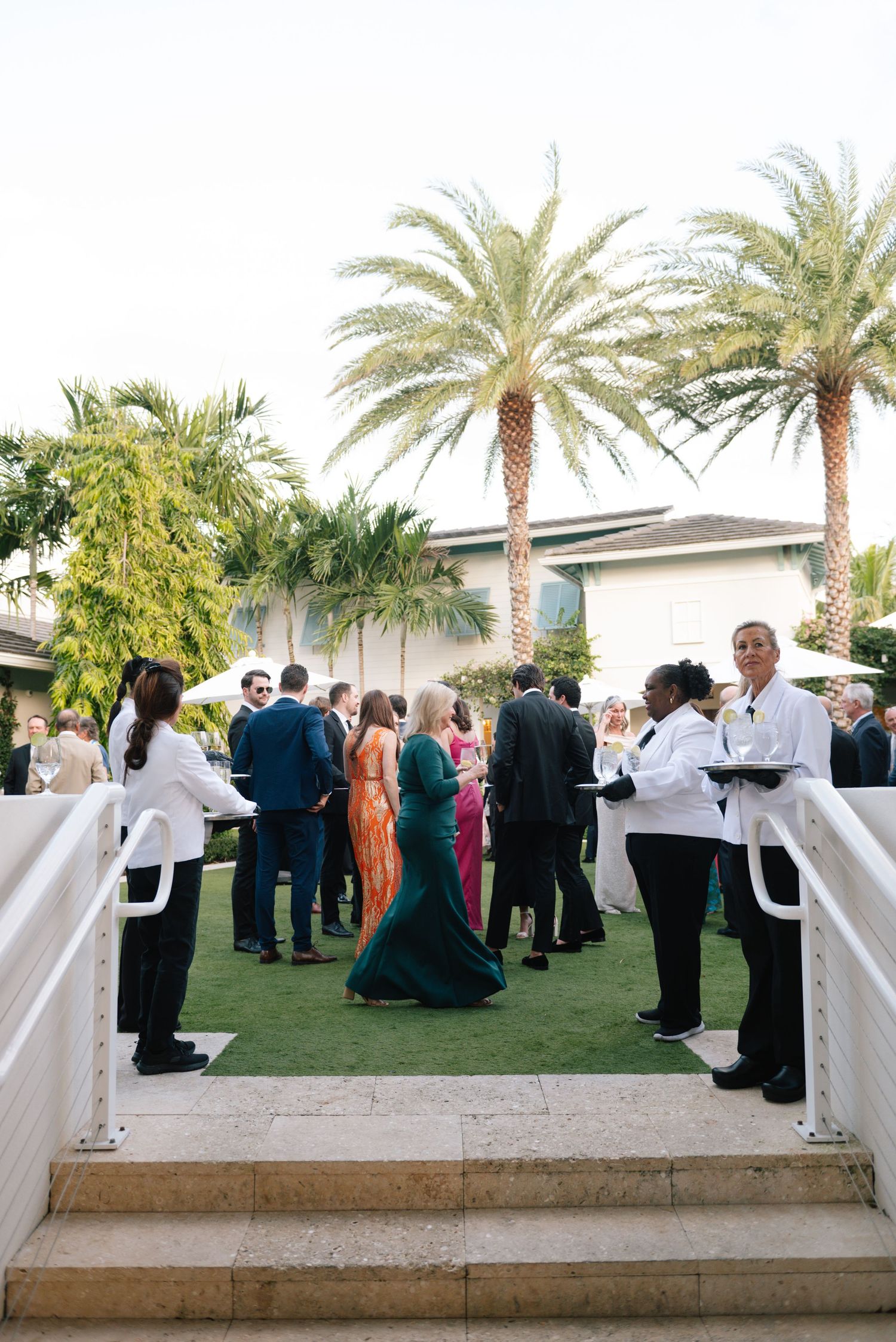 Wedding reception guests mingle on lawn with palm trees at outdoor venue.