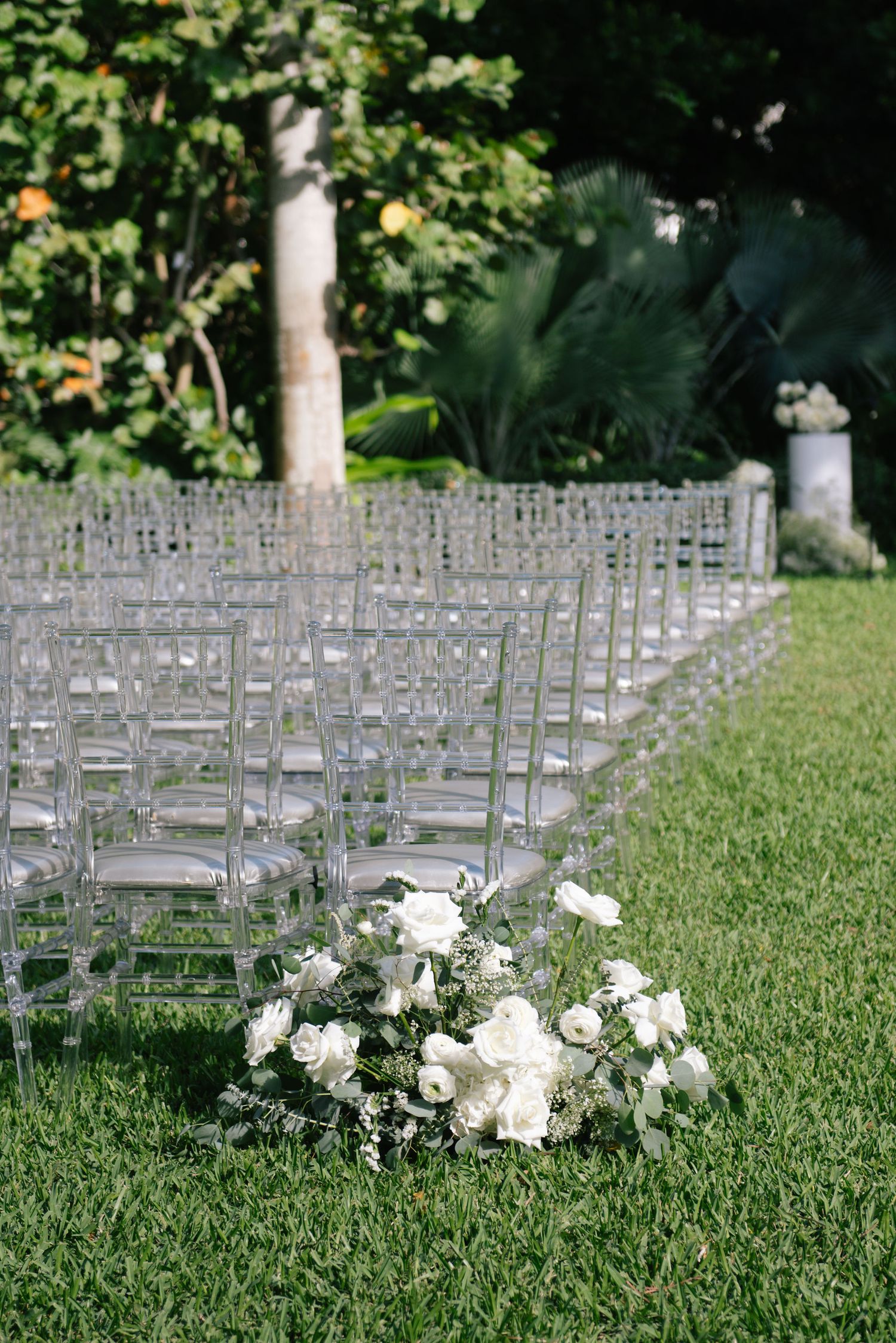 Rows of clear acrylic chairs set up for an outdoor wedding ceremony with white floral arrangements on the grass.