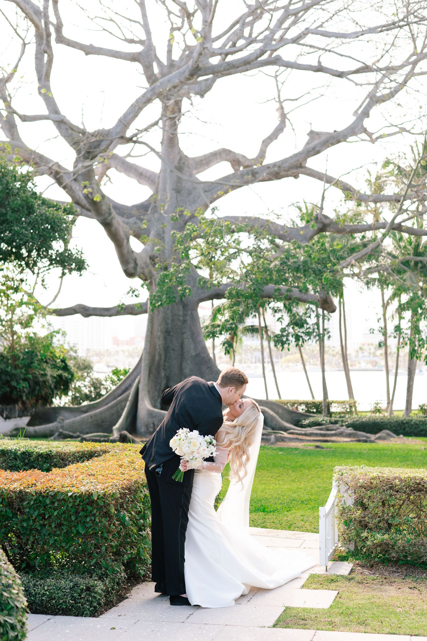 Wedding couple poses romantically under a sprawling banyan tree in a garden setting.