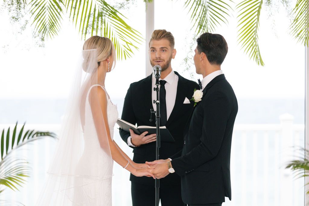Wedding ceremony in a bright indoor venue decorated with tropical palm leaves and white flowers.