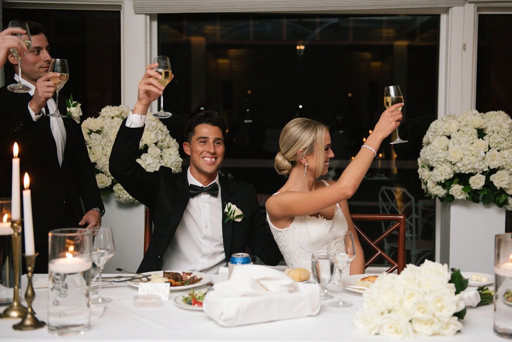 Wedding guests raise their glasses in a celebratory toast at a reception with white floral decorations.