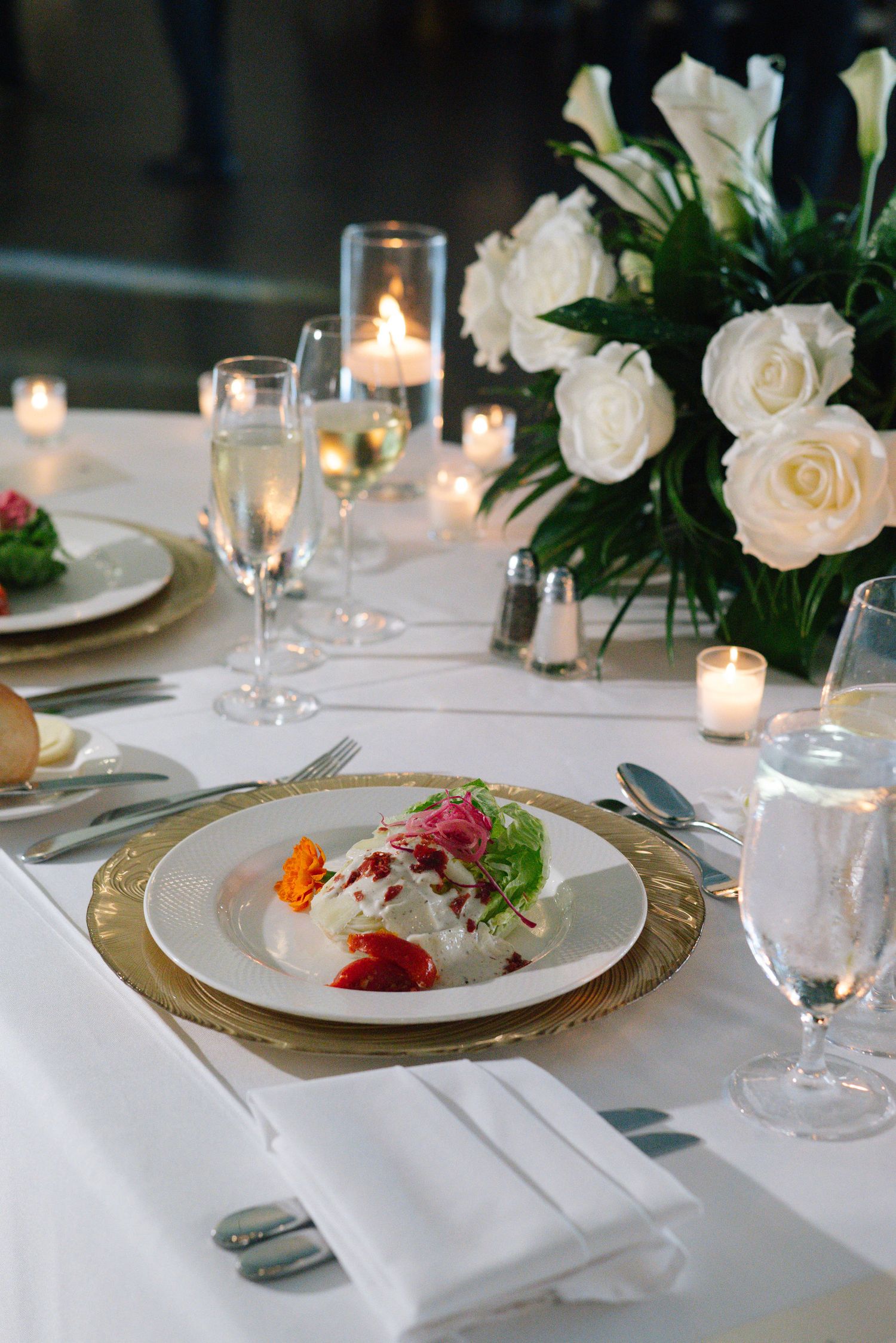 Elegant place settings with gold chargers, white roses, and candlelight on crisp white tablecloths.