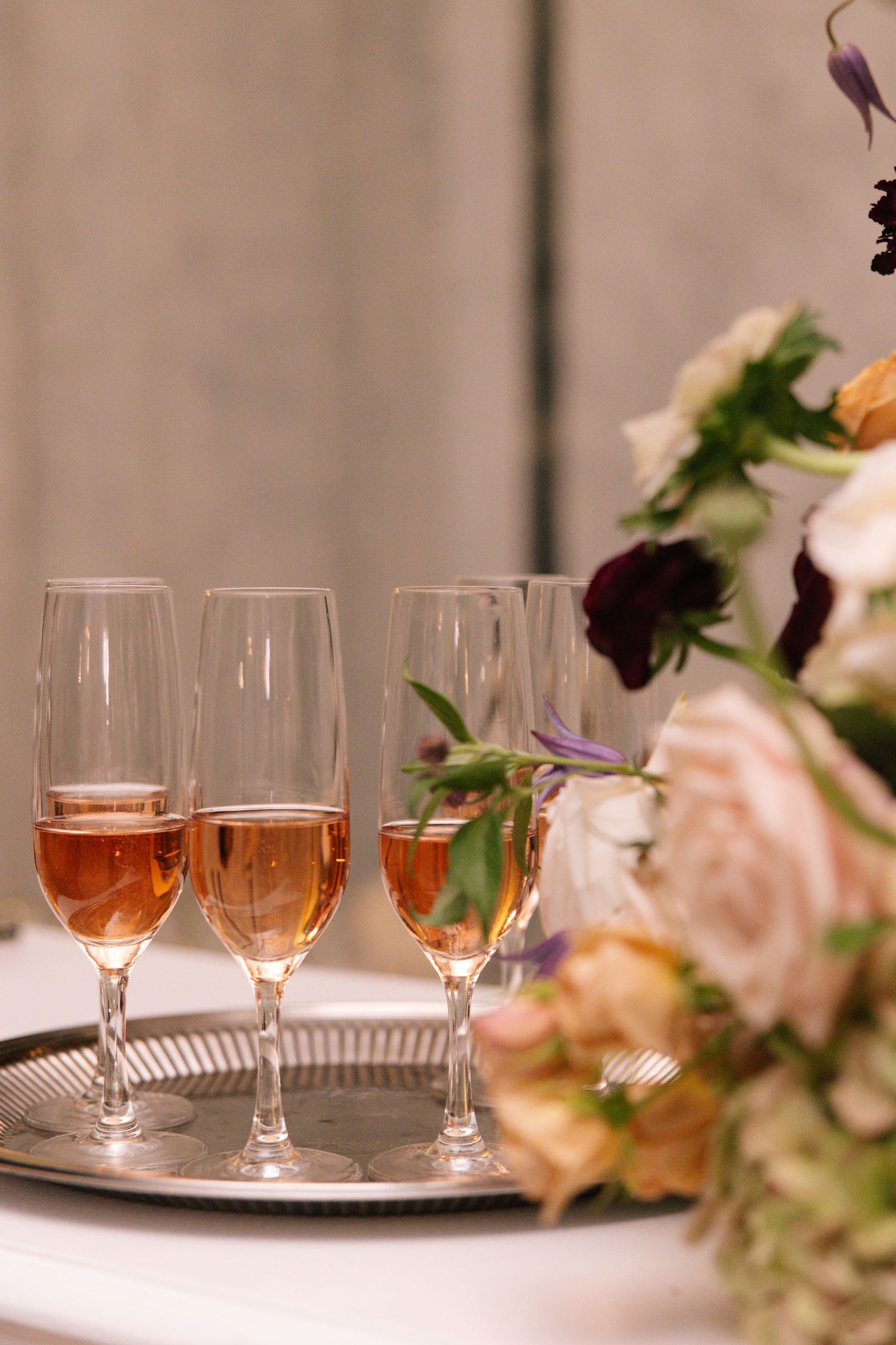 Three champagne flutes filled with rose wine on silver tray next to floral arrangement.