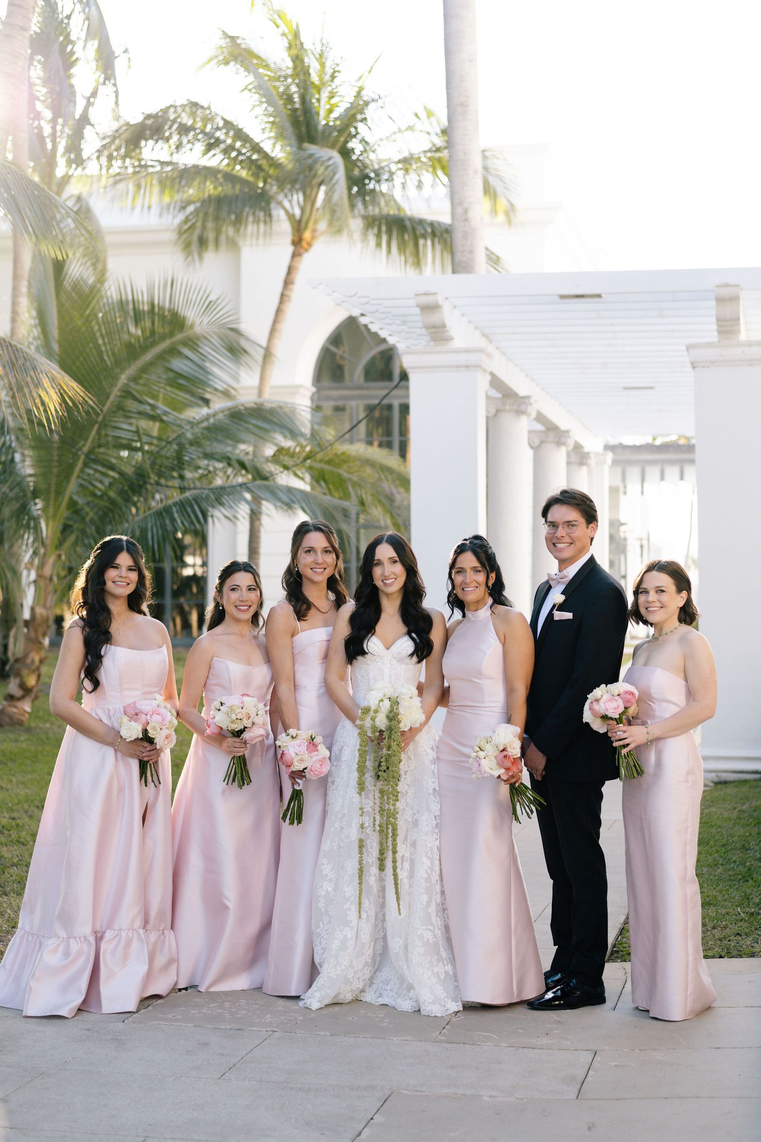 Wedding party in pink bridesmaid dresses poses by white columns and palm trees.