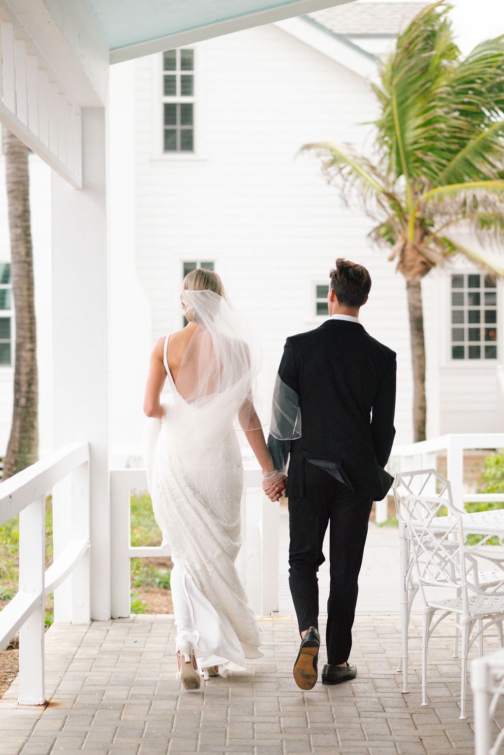Newlyweds walking along white porch with palm trees in tropical wedding setting.