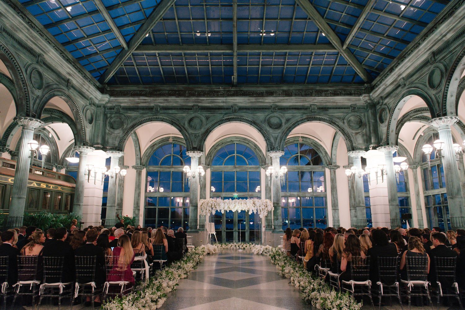 Stunning wedding ceremony setup in glass-ceiling venue with rows of white chairs and floral decorations.