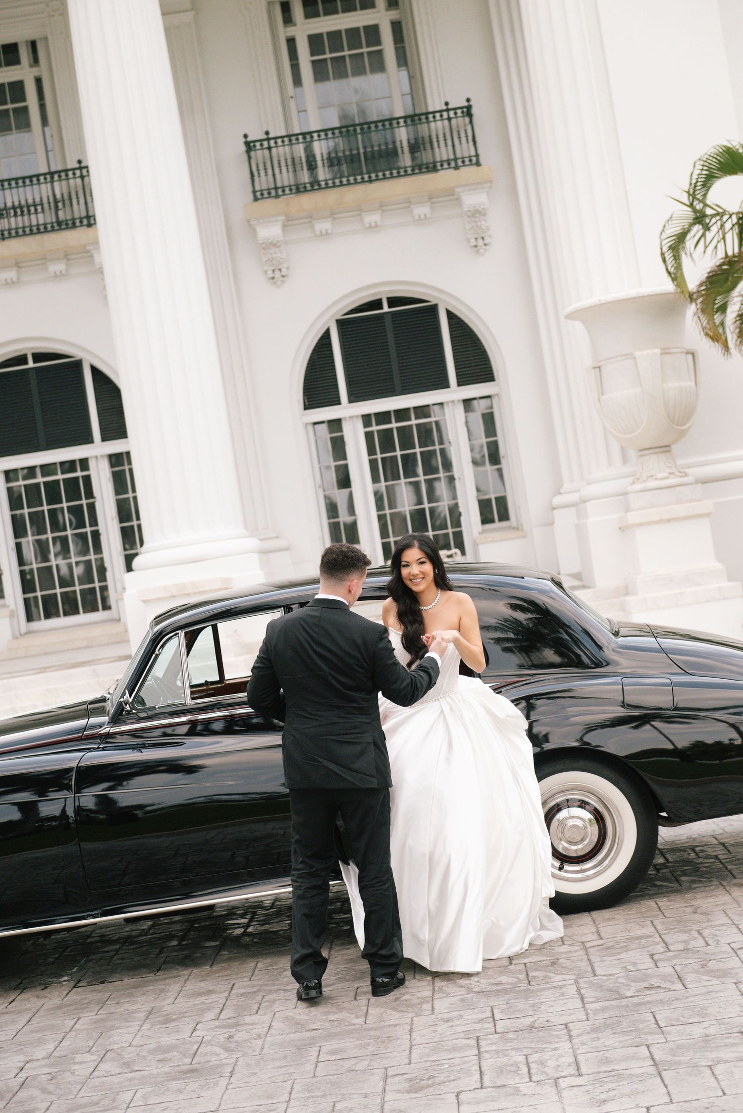 Couple in wedding attire near vintage black car outside white colonial building.