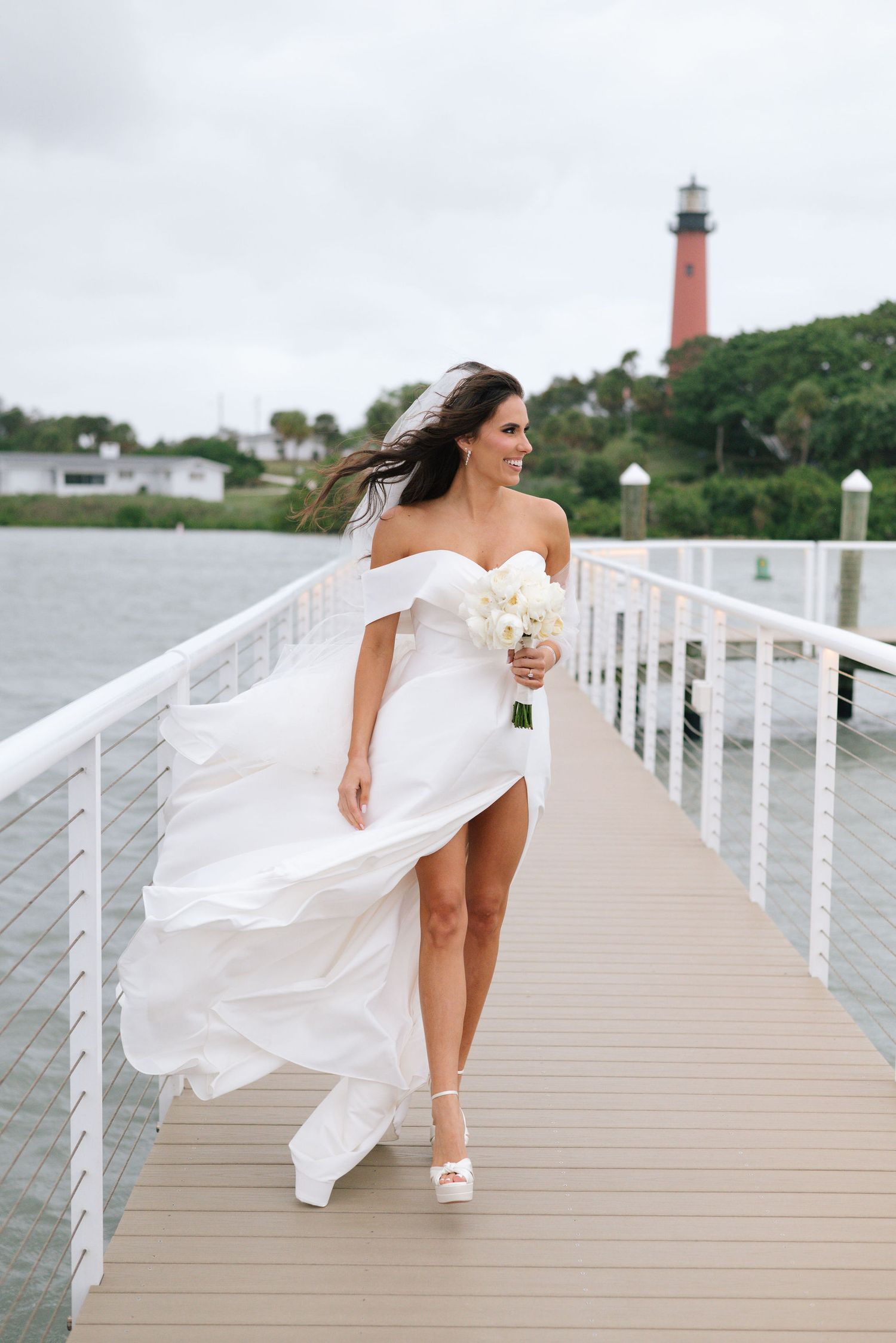 A white wedding dress flows in the breeze on a waterfront boardwalk with a lighthouse in the background.