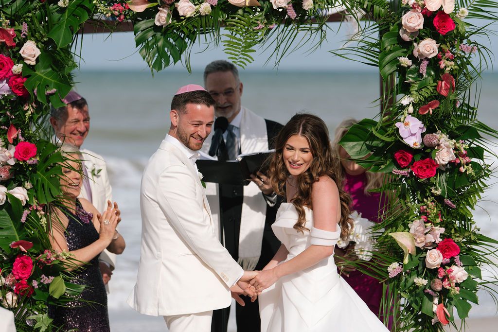 Celebration moments during beach wedding under floral arch with pink and white flowers.