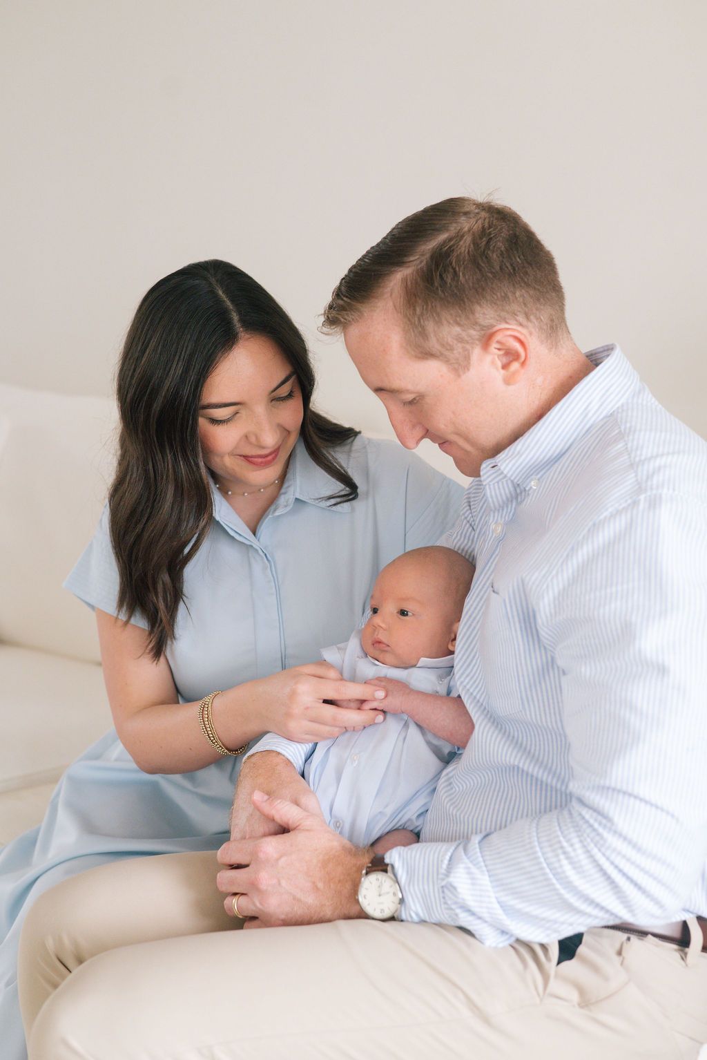 Parents in matching blue and white attire share tender moment with their newborn on white couch.
