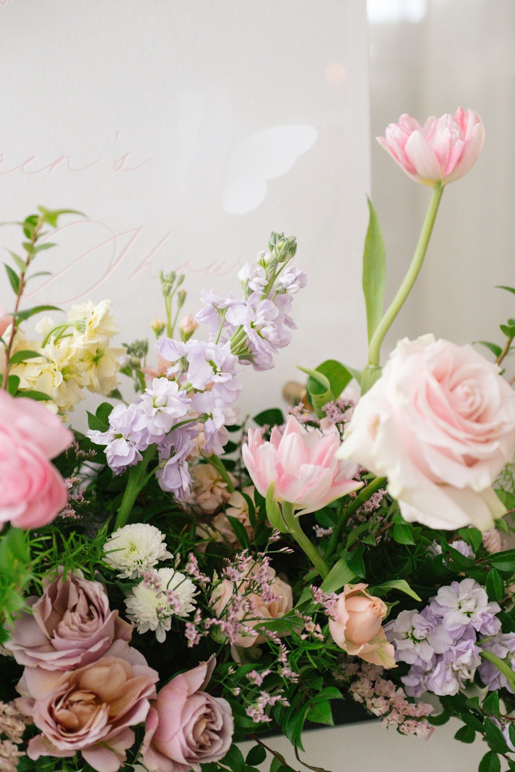 Close-up of lush floral arrangement featuring pink roses, tulips, and purple blooms against white background.