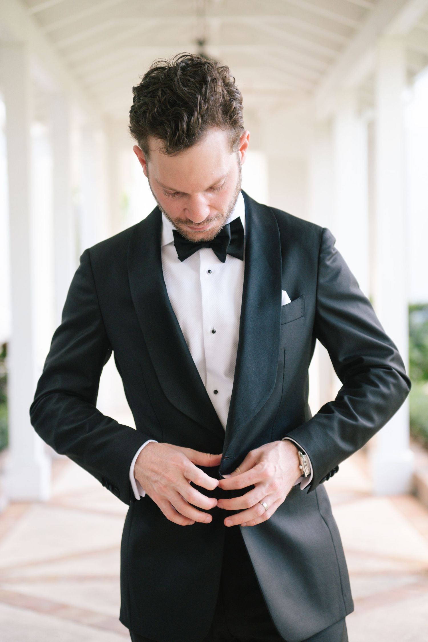Groom adjusting his black tuxedo jacket in an elegant white hallway.