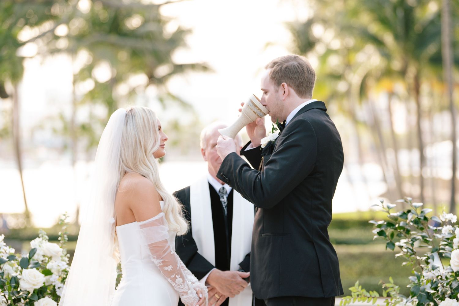 A couple exchanges vows during an outdoor wedding ceremony with palm trees and white decor in the background.