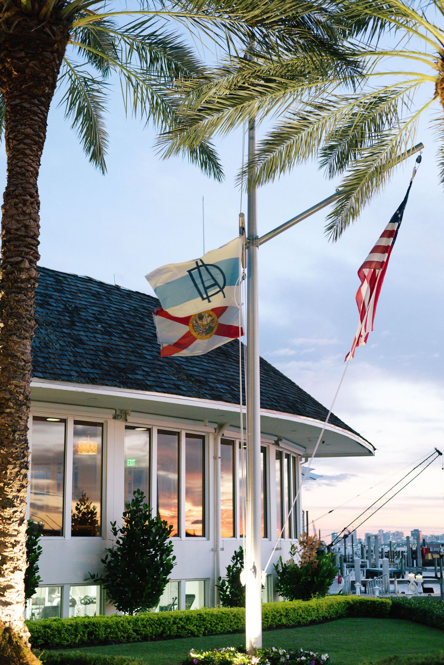 Coastal yacht club building with American flag and palm trees at sunset.