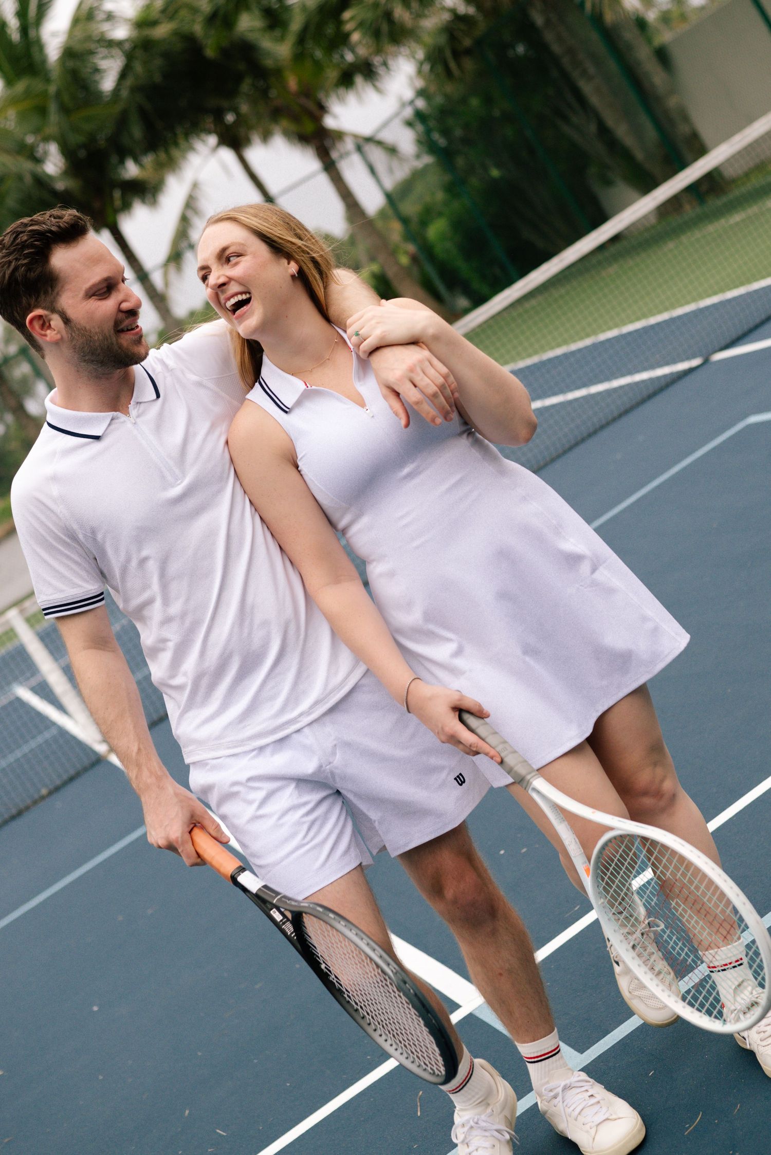 Two people in matching white tennis outfits share a playful moment on the court.