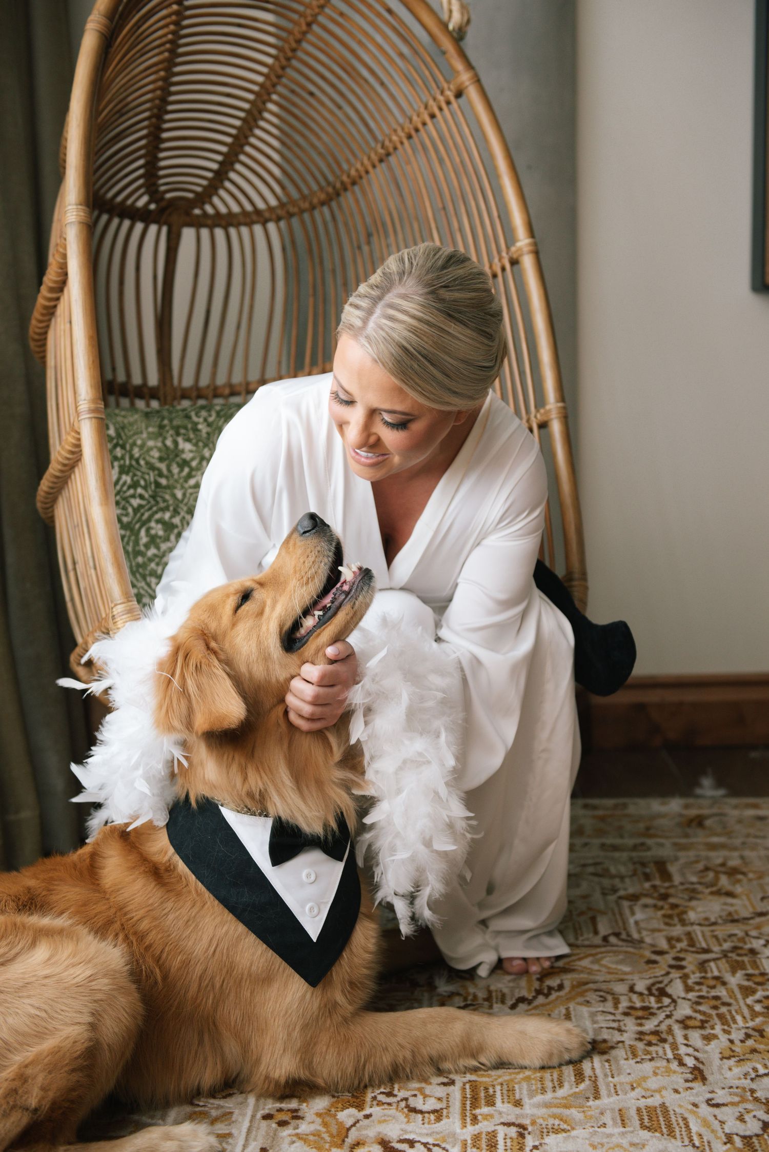 Bride in white robe kneels beside golden retriever wearing tuxedo and feather boa near rattan egg chair.