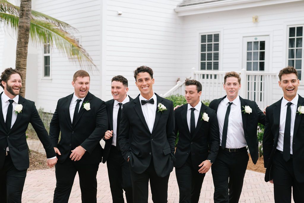 Large wedding party in formal attire poses together outside white coastal venue.