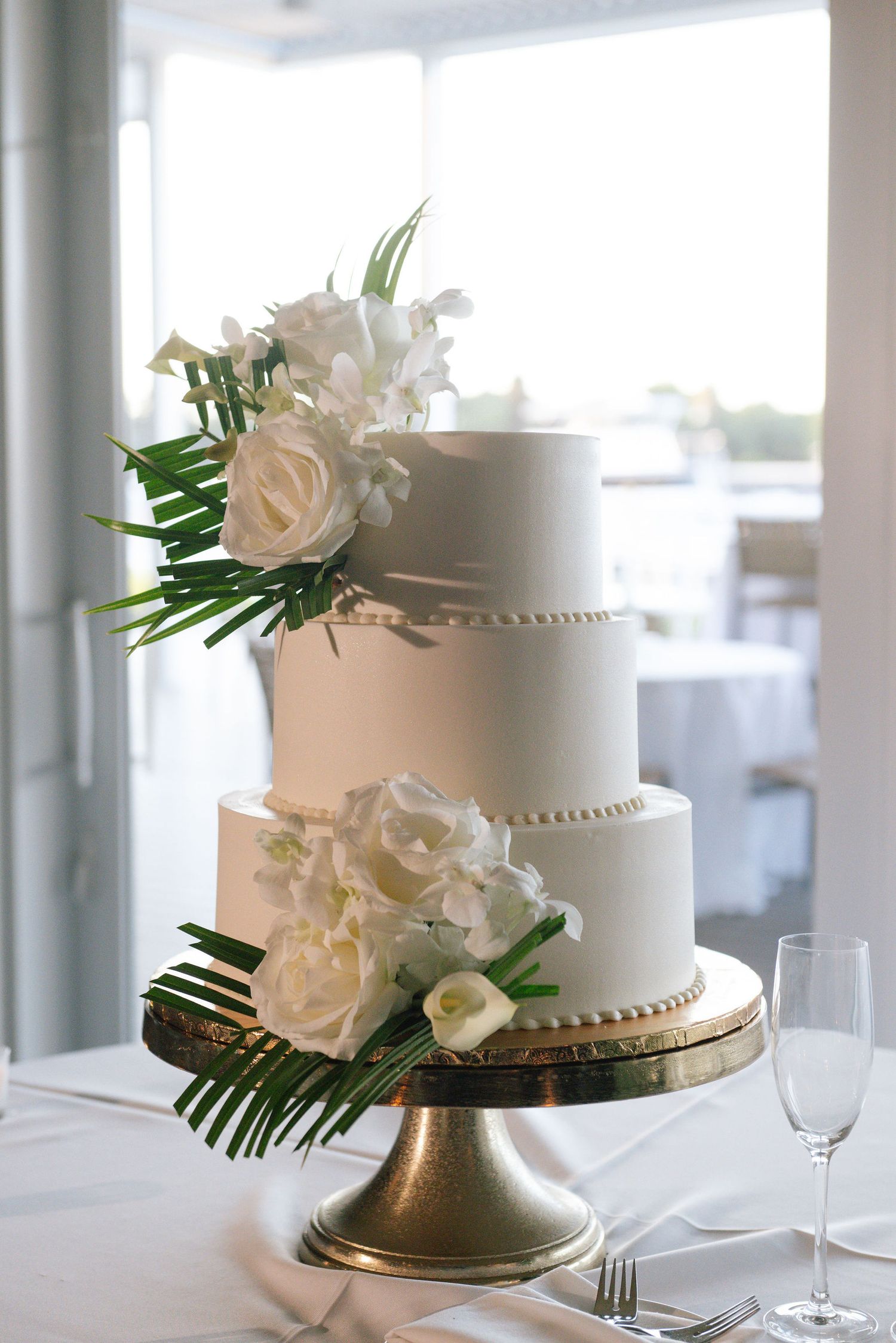 Classic three-tier white wedding cake decorated with fresh roses and palm leaves on a gold stand.