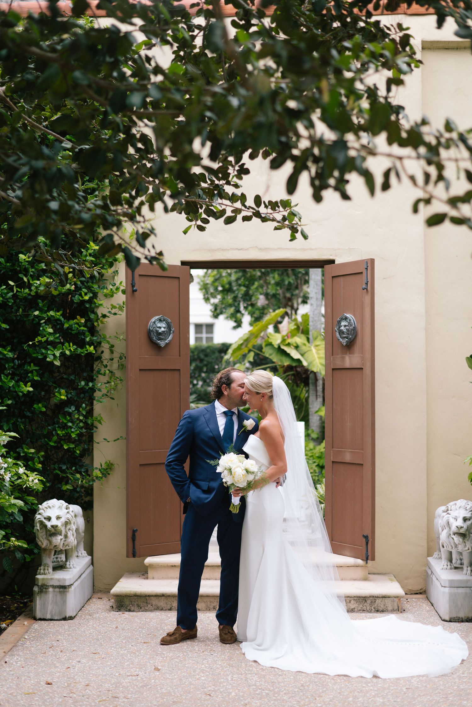 Wedding couple posing by rustic brown doors in a Mediterranean-style courtyard.