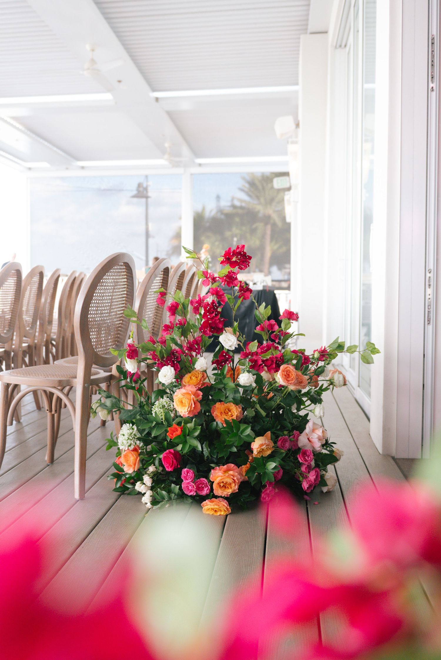 Bright floral arrangements with pink and orange blooms decorate a sunlit porch with brown wicker chairs.