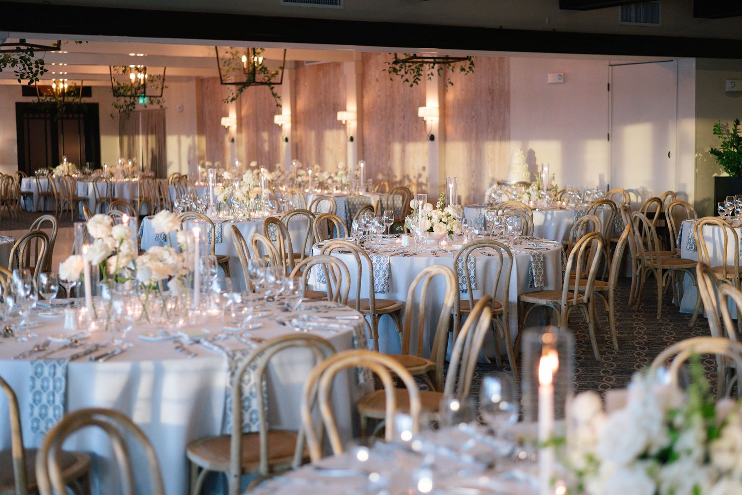 Elegant wedding reception tables set with white linens, glassware and floral centerpieces in a bright ballroom.