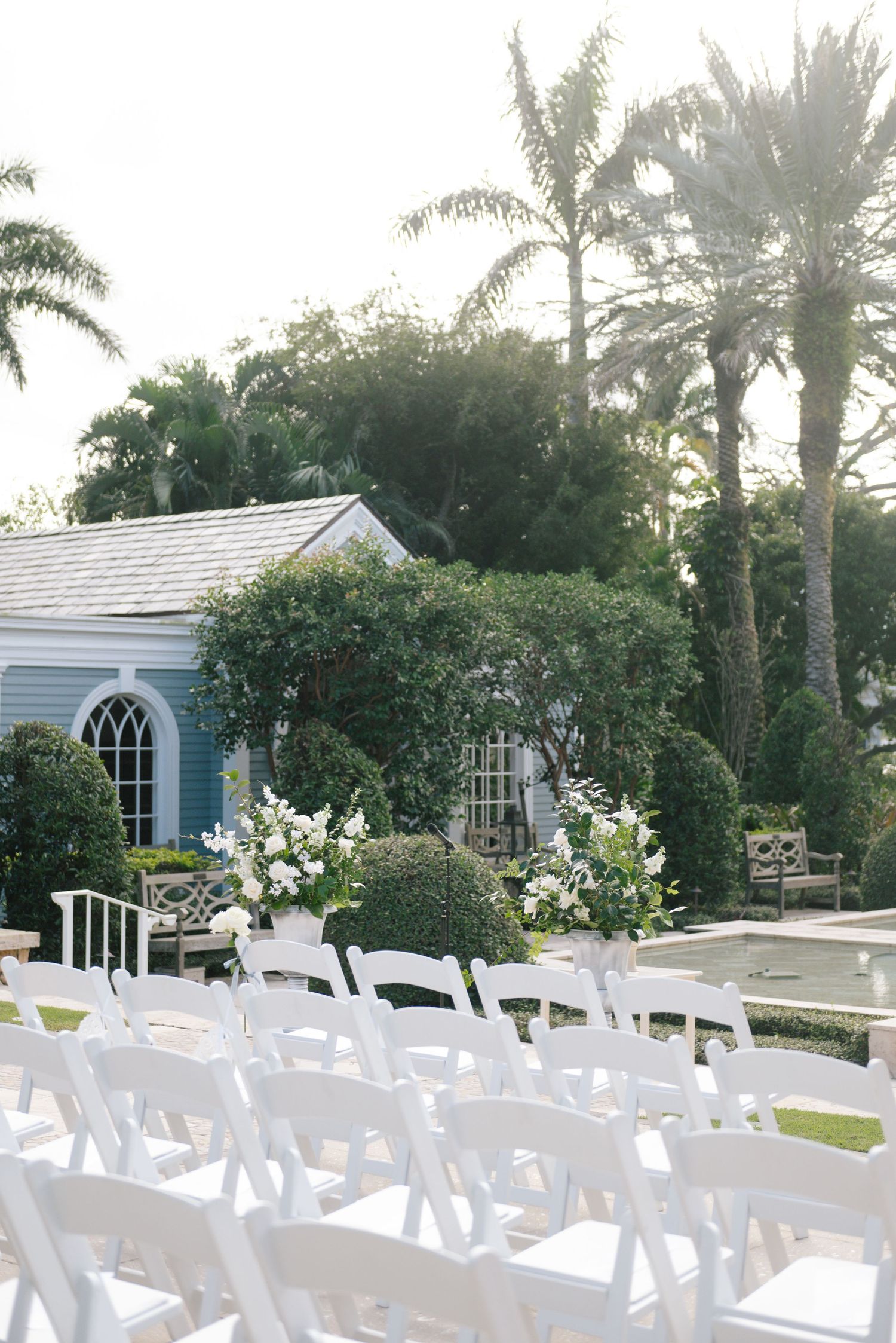 A serene outdoor wedding venue setup with white chairs and palm trees against a sunset sky.