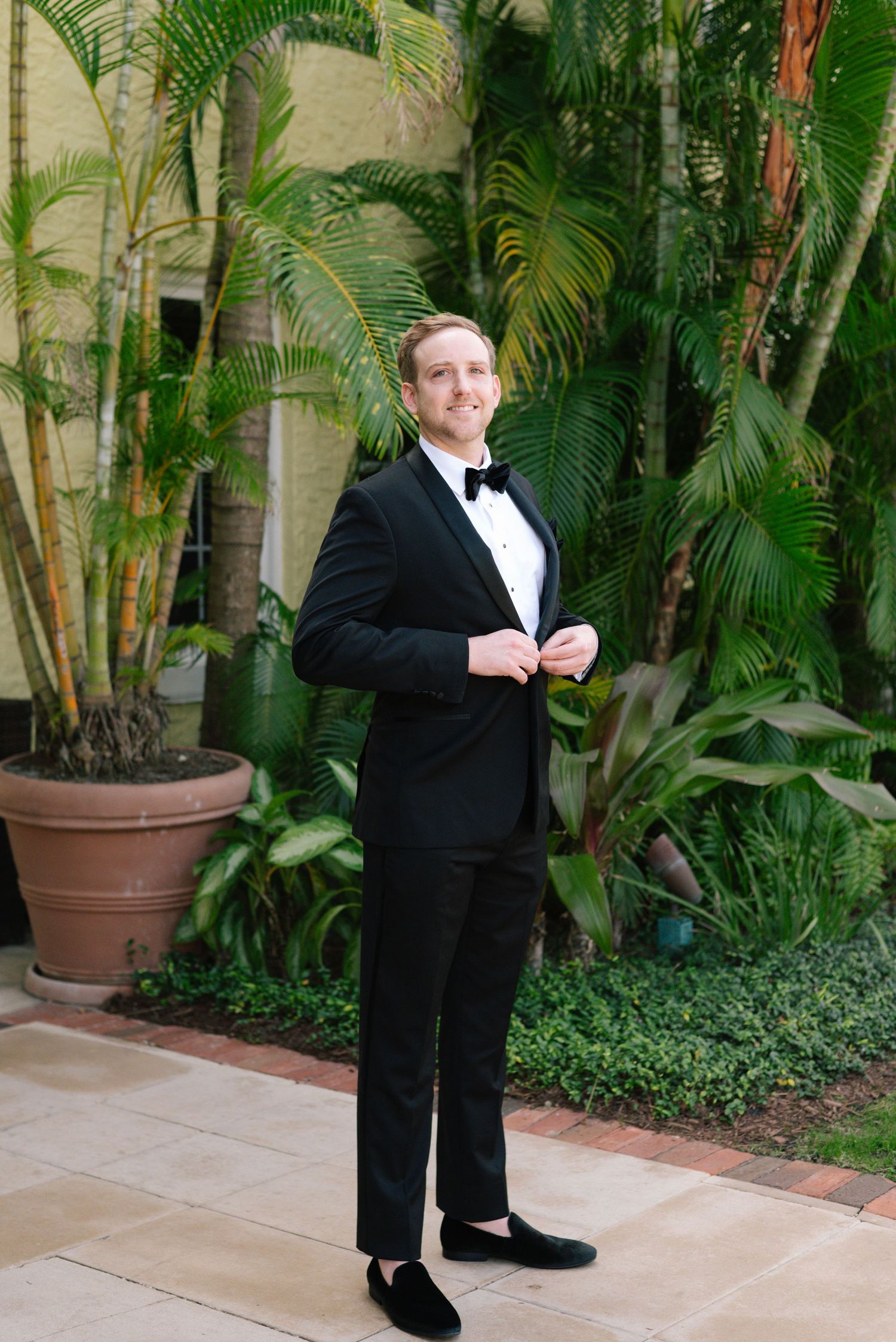 Groom in black tuxedo poses playfully in front of tropical garden setting with palm trees.