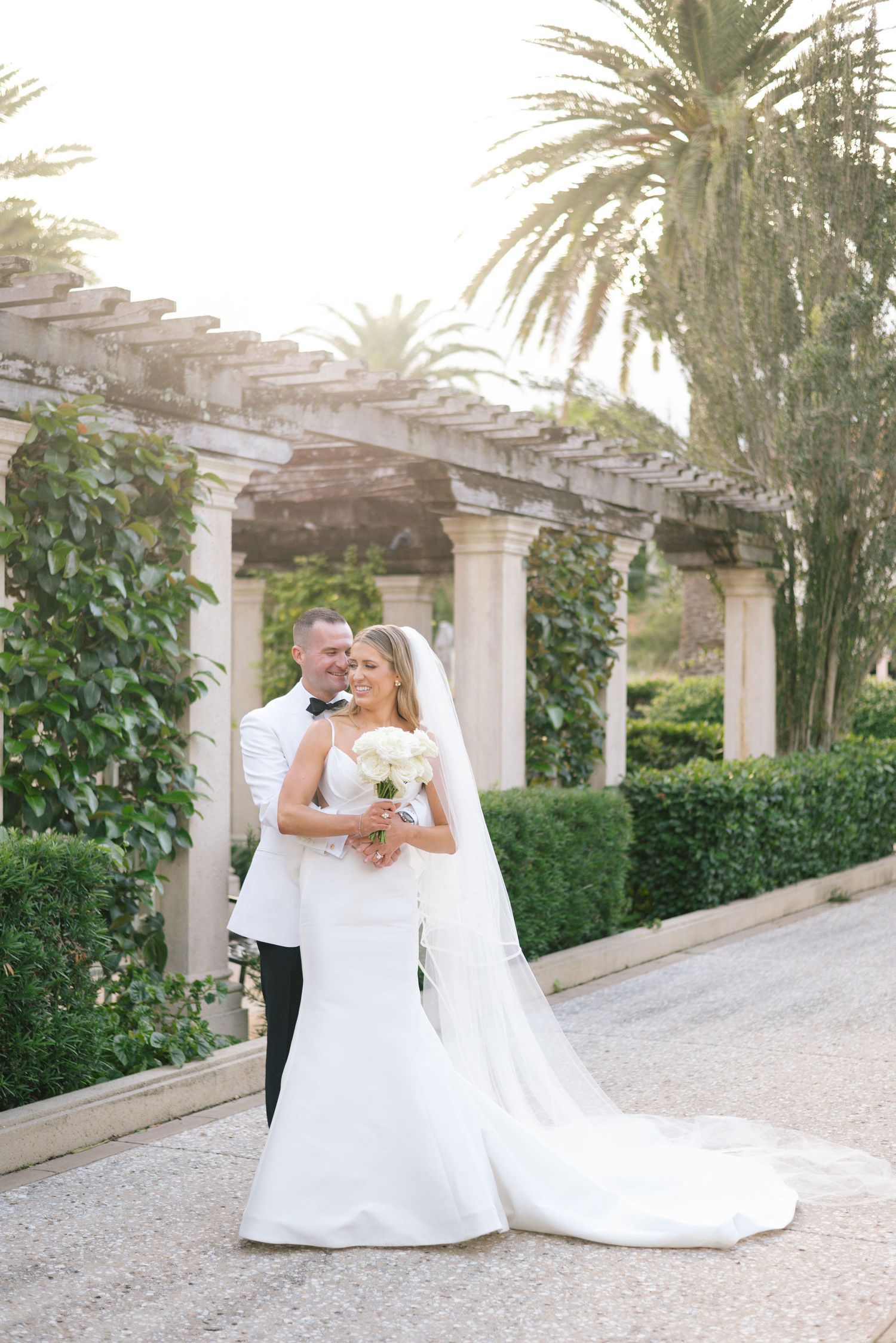 A couple shares an intimate moment under a vine-covered pergola with palm trees in the background.