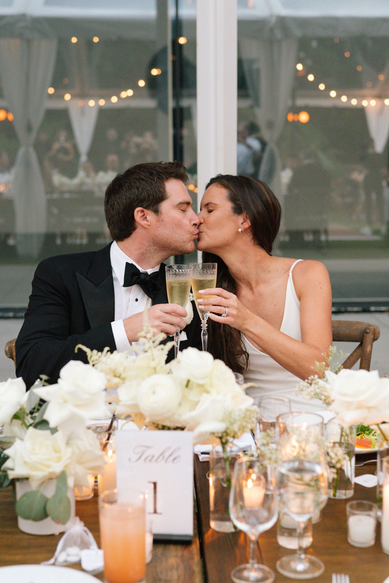 Newlyweds share a romantic toast at their reception table decorated with white flowers and string lights.