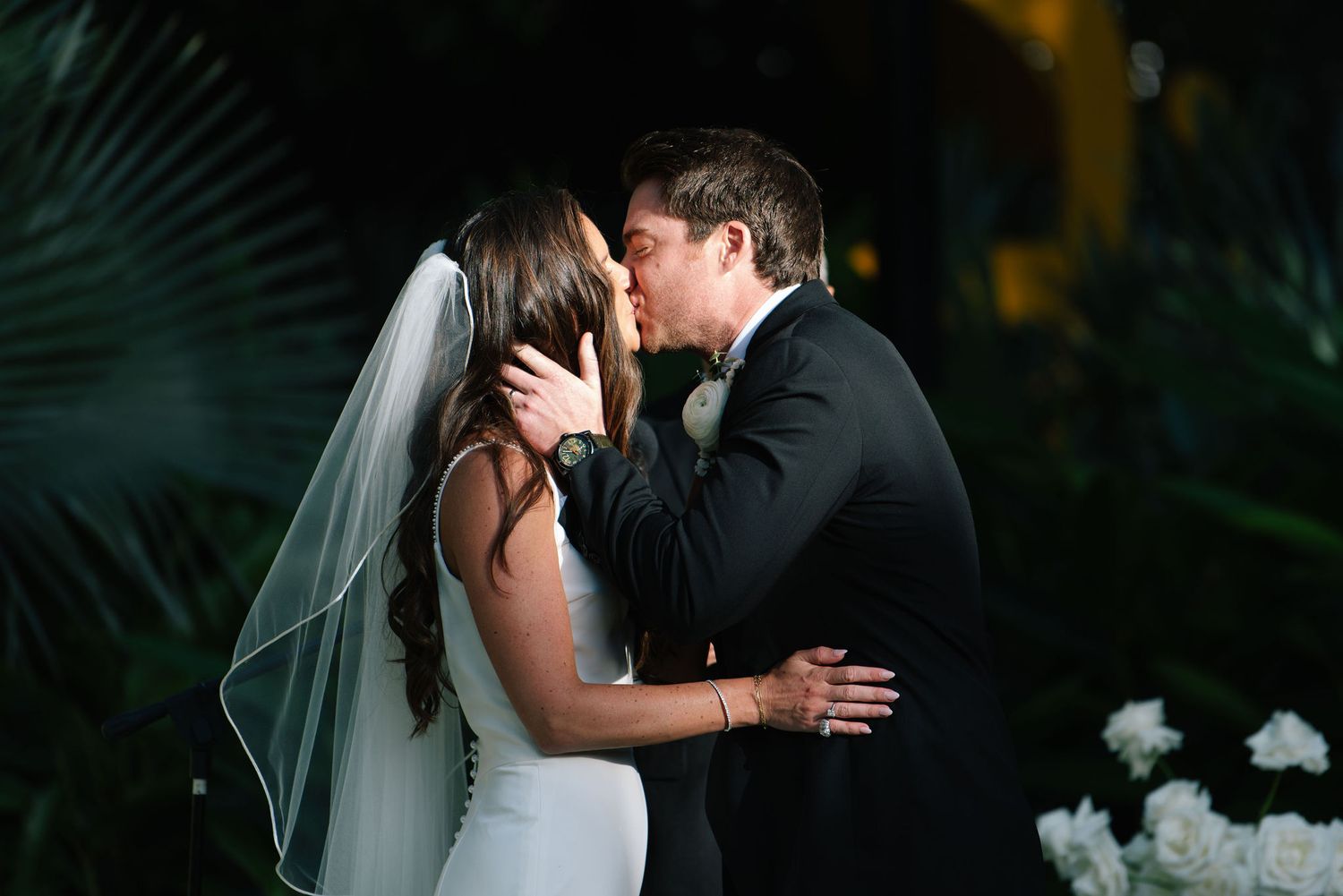 Romantic wedding kiss moment against a backdrop of tropical palm fronds at dusk.