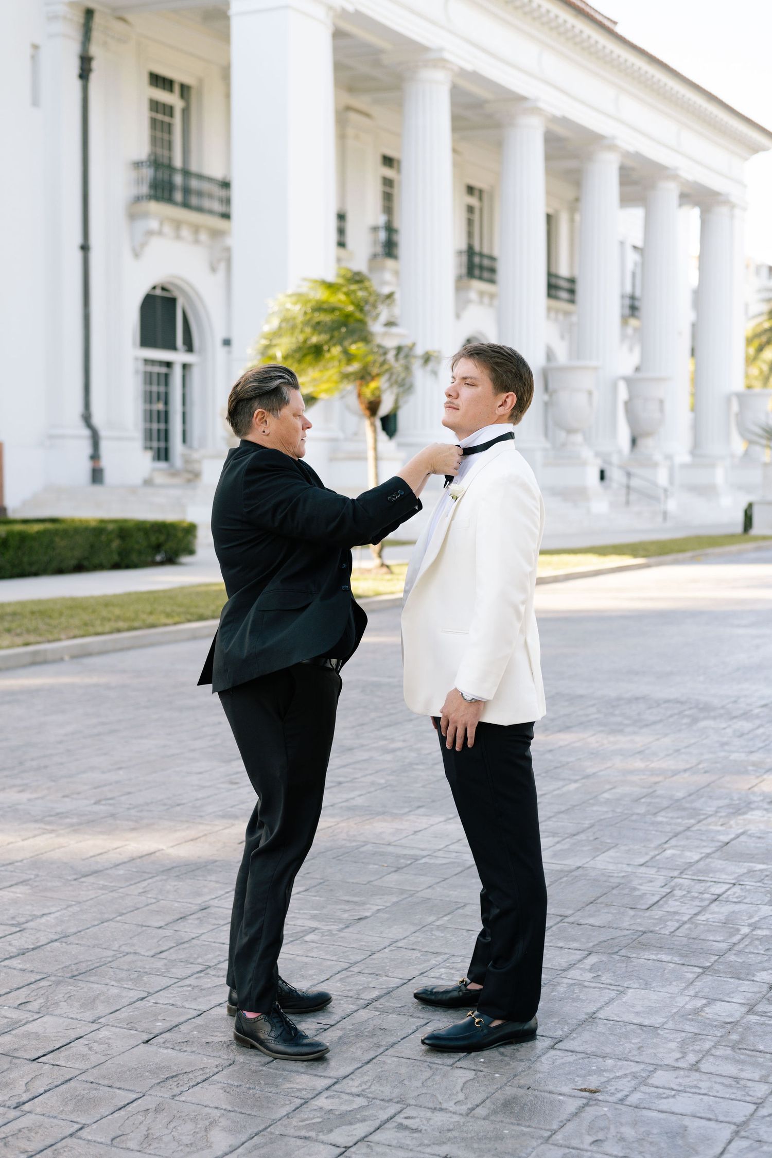 Two figures in formal attire interacting on a stone walkway in front of white columns.