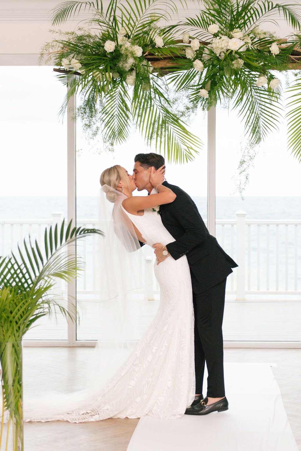 First kiss as newlyweds under elegant palm and white floral arch.