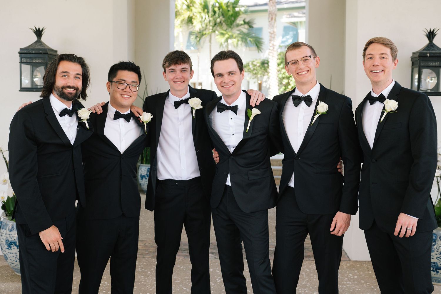 Groomsmen in black tuxedos pose playfully in a line at an outdoor wedding venue.