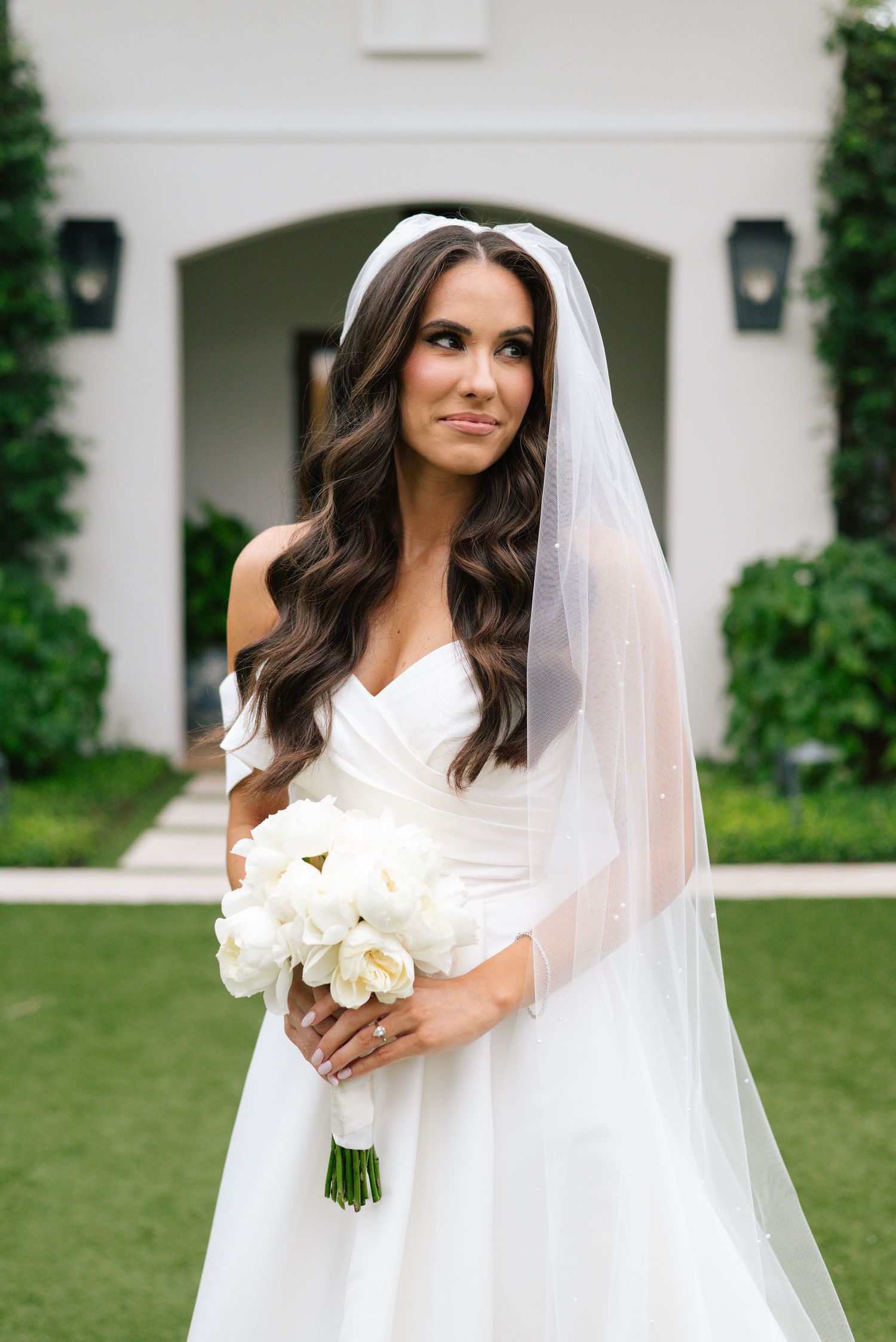 Bride in classic white gown holds white rose bouquet against architectural backdrop.