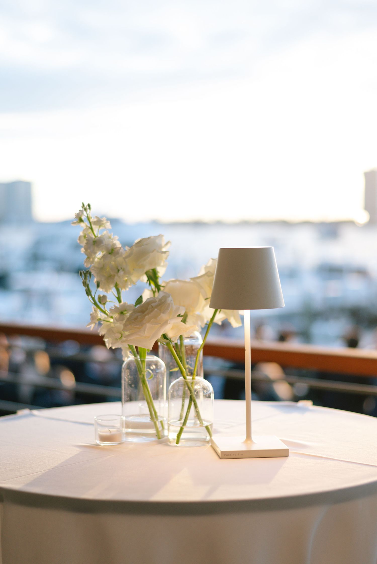 White floral centerpieces and table lamps create an elegant dining atmosphere with a city view in the background.