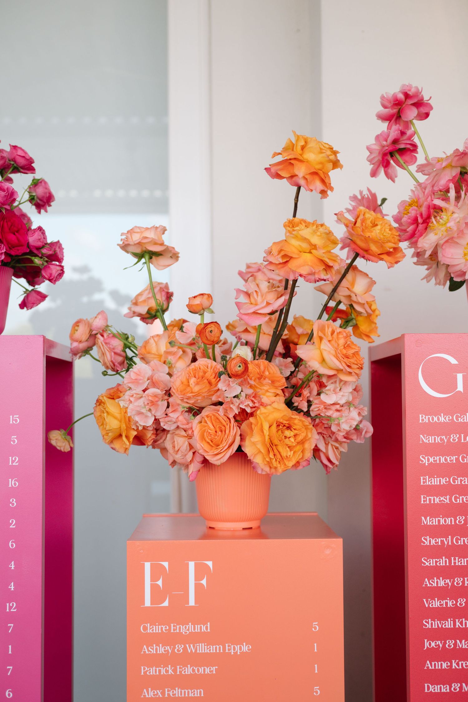 Wedding seating chart display with coral and pink floral arrangements against a white wall.